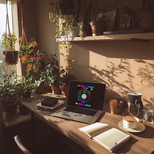 image of the product for sale of a colored hex map is shown on an open laptop on a desk in a sun filled room with plants, an open notebook, coffee, and general coziness